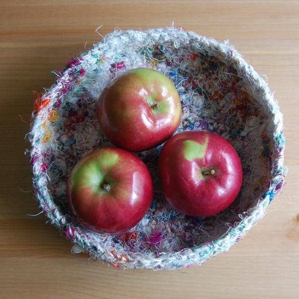 A birds eye view of a white tray made of multicolored white yarn sitting a wooden table with apples inside of the tray