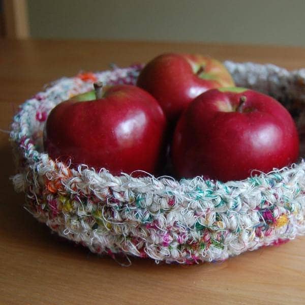A white tray made of multicolored white yarn sitting a wooden table with apples inside of the tray