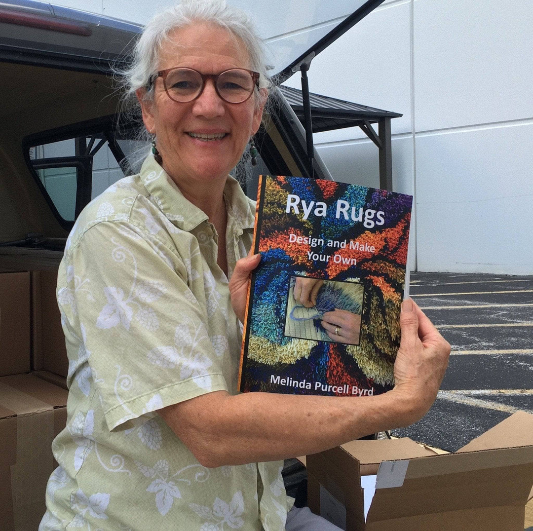 Melinda Purcell Byrd holding her book "Rya Rugs" in front of a car, in a parking lot, with cardboard boxes around.