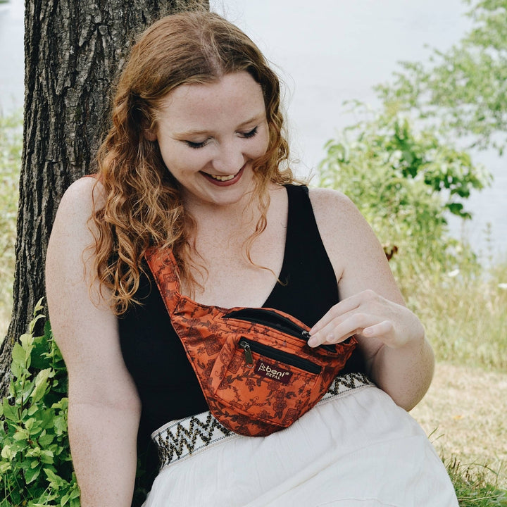 Model is sitting by a tree wearing an orange sari silk fanny pack across their body. 