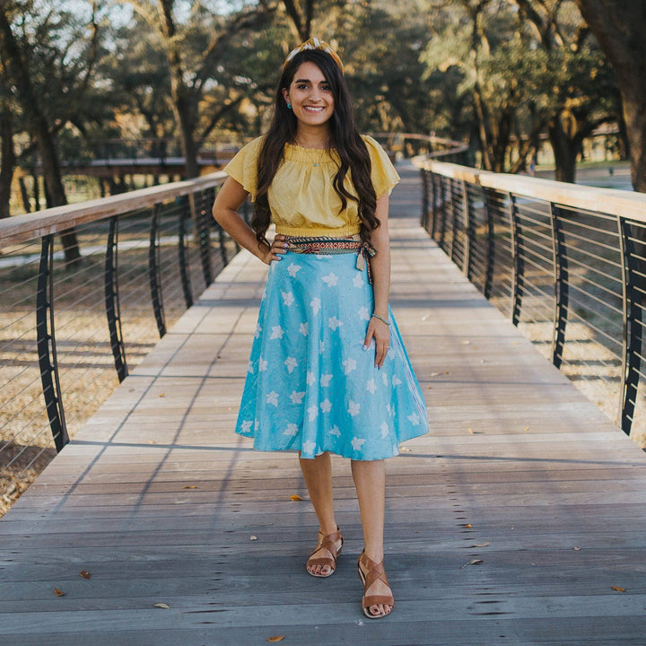 A girl standing on a board walk wearing a baby blue sari wrap skirt with white orchids on it. She's paired it with a light yellow crop top and a kameela knot sari silk headband.