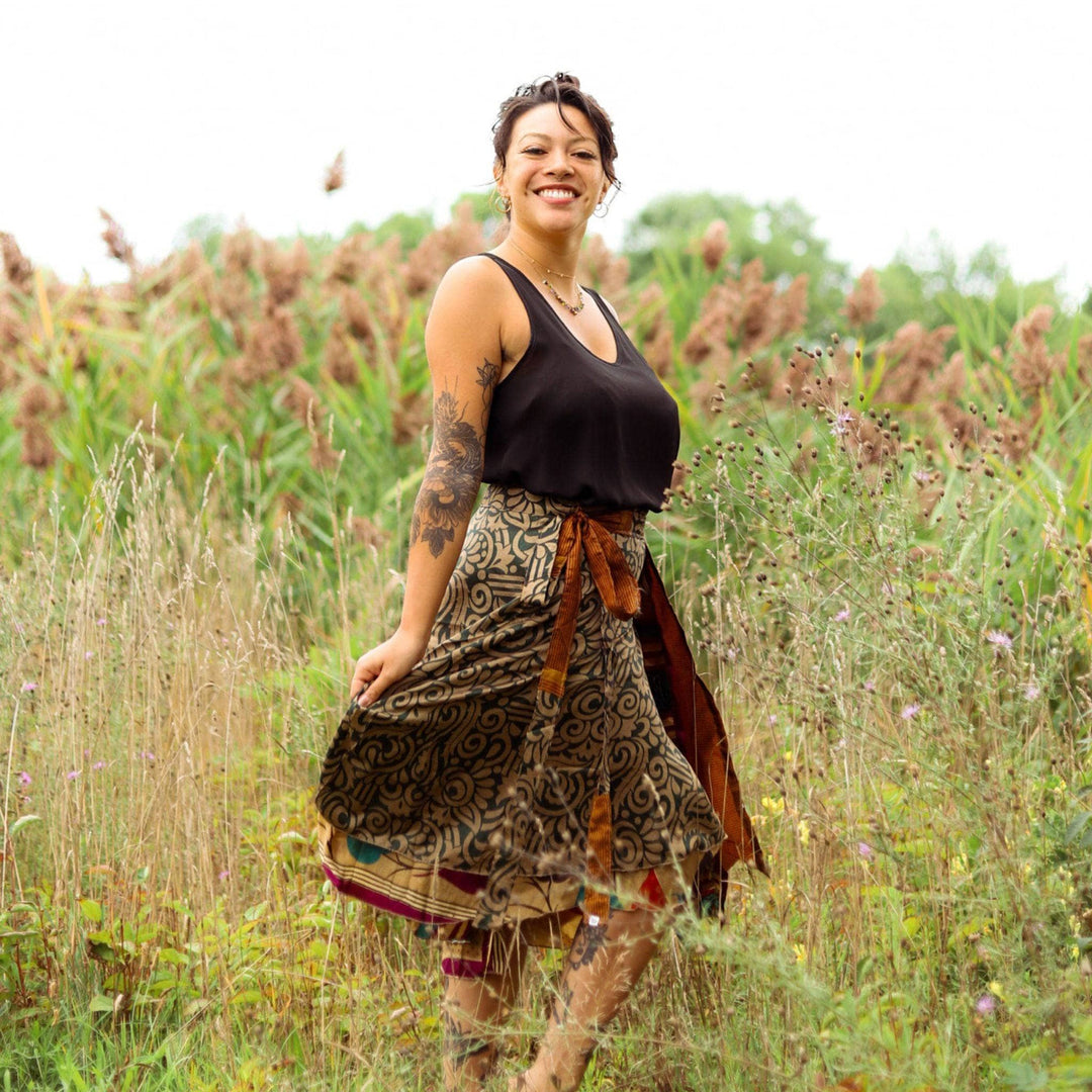 A girl standing in a field wearing a tea length sari wrap skirt in warmer tones. The Skirt is gold with darn green accents all over. She's wearing a black tank top with it and gold jewelry.