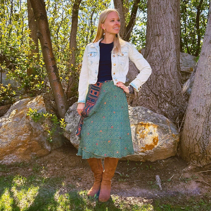 A girl standing in a forest wearing an emerald green paisley tea length skirt made from reclaimed sari material. The Under layer is a navy and light pink combo. She's paired the skirt with a tall brown Swede boot, a navy blue shirt and a white washed denim jacket. 