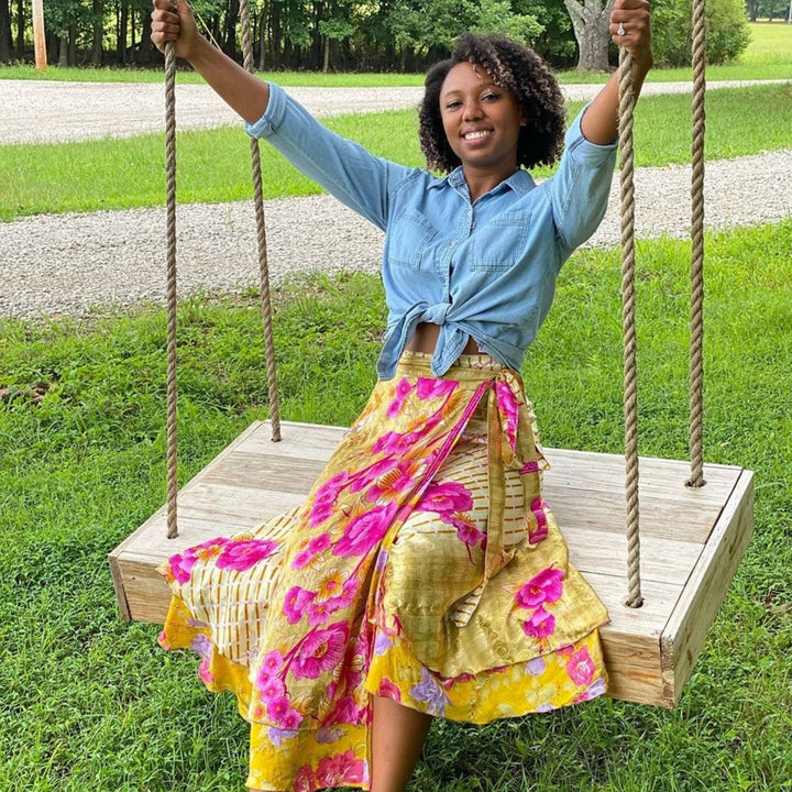 A girl sitting on a swing wearing a gold maxi length sari wrap skirt with light pink flowers all over it. She's wearing a cropped denim long sleeve shirt that's tied.