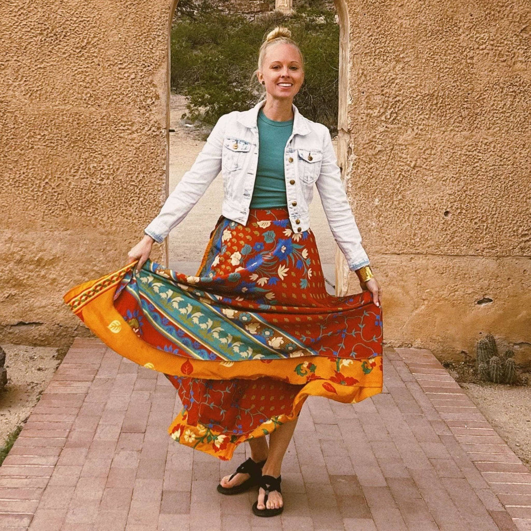 A woman standing in front of a clay stone building in the dessert. She's wearing a maxi skirt made from reclaimed sari. The skirt is maroon with green and gold stripes and blue flowers all over it. She's wearing a light green shirt with it and a light wash denim jacket overtop.