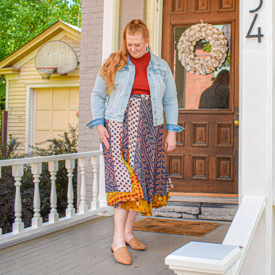 A woman walking off of her front porch. She's wearing a tea length wrap skirt mad from reclaimed sari. The top layer is navy blue with leaf patterns all over it while the second under layer is gold with light yellow circles. SHe's wearing a read sweater with it and a light denim jacket.