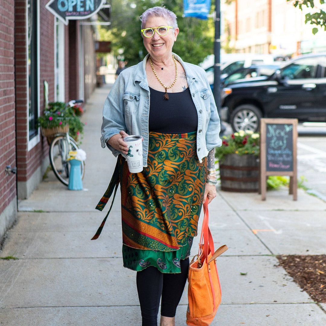 A woman walking down the street wearing gold and green sari wrap skirt with black leggins and a black tank top. She's walking down the street with a cup of coffee.