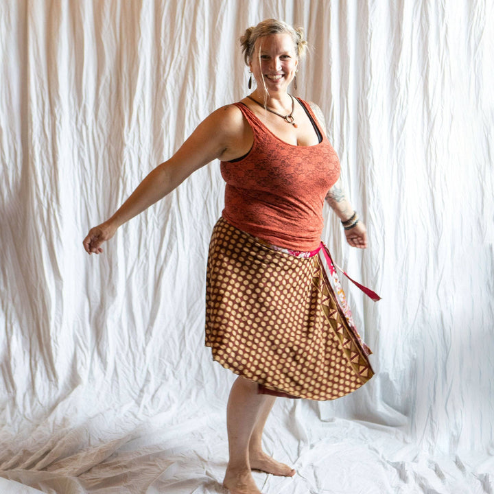 A woman standing in front of a photo backdrop wearing a brown sari wrap skirt with gold dots all over it. She's paired it with a burnt orange tank top and chunky jewelry.