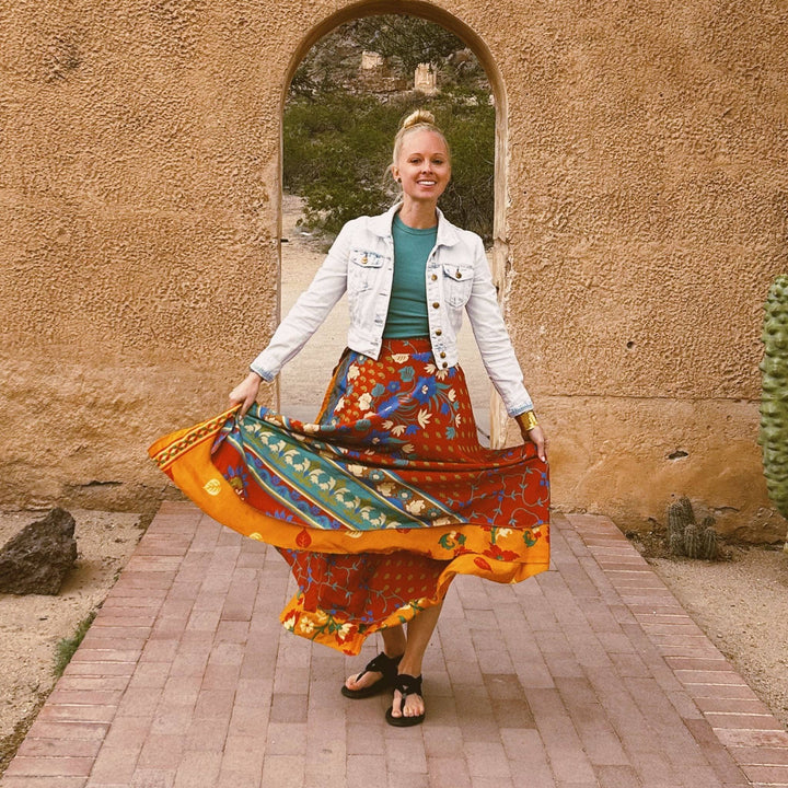 A woman standing in front of a clay wall with a burnt read sari wrap skirt on. The top layer is burnt red while the bottom layer is bright orange. There's a colorful leaf pattern on both layers