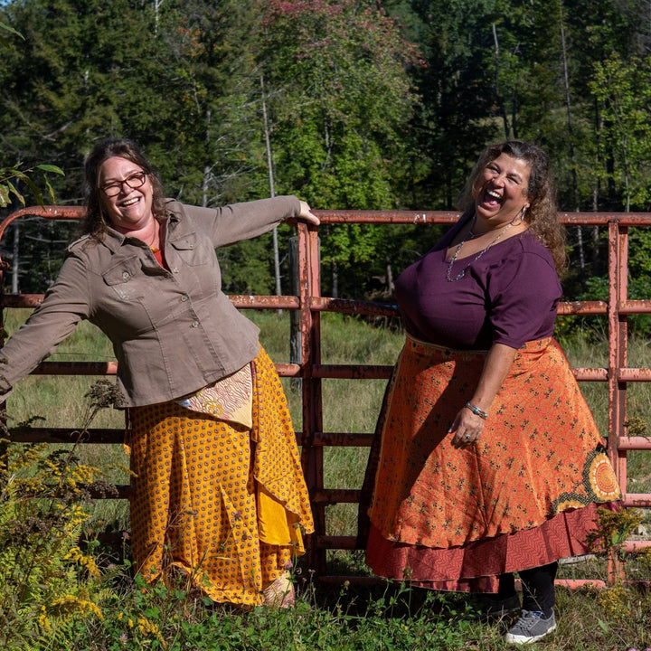 Two models laughing wearing warm toned sari wrap skirts in a farm field. 