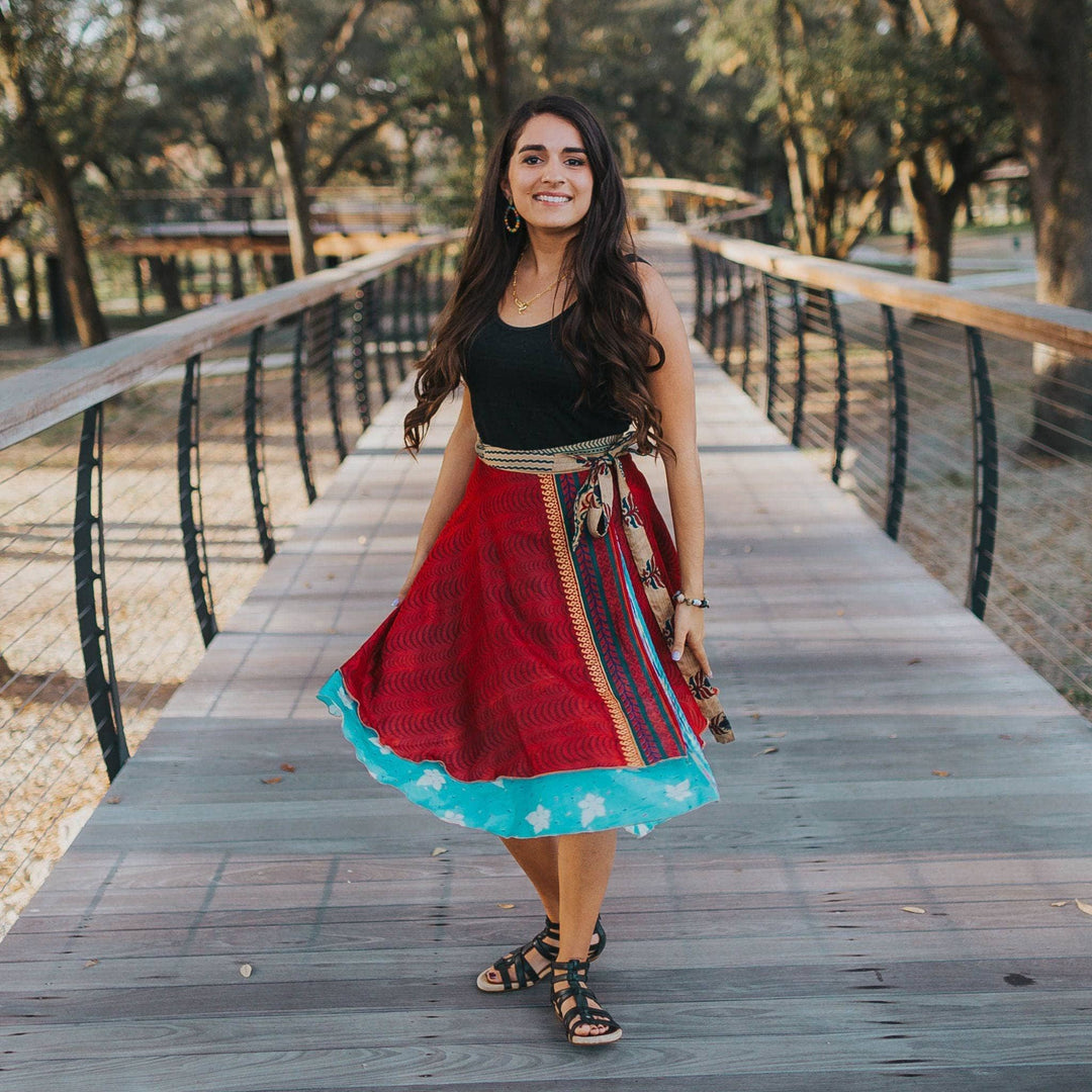 Model wearing a Size 00-04 Sari Wrap Skirt in Warmer Tones. The skirt is Tea Length. The top layer is a rich maroon color with zig zag patterns and tan accents, the under layer is a bright blue. She's paired the skirt with a simple black tank top and gladiator sandal.