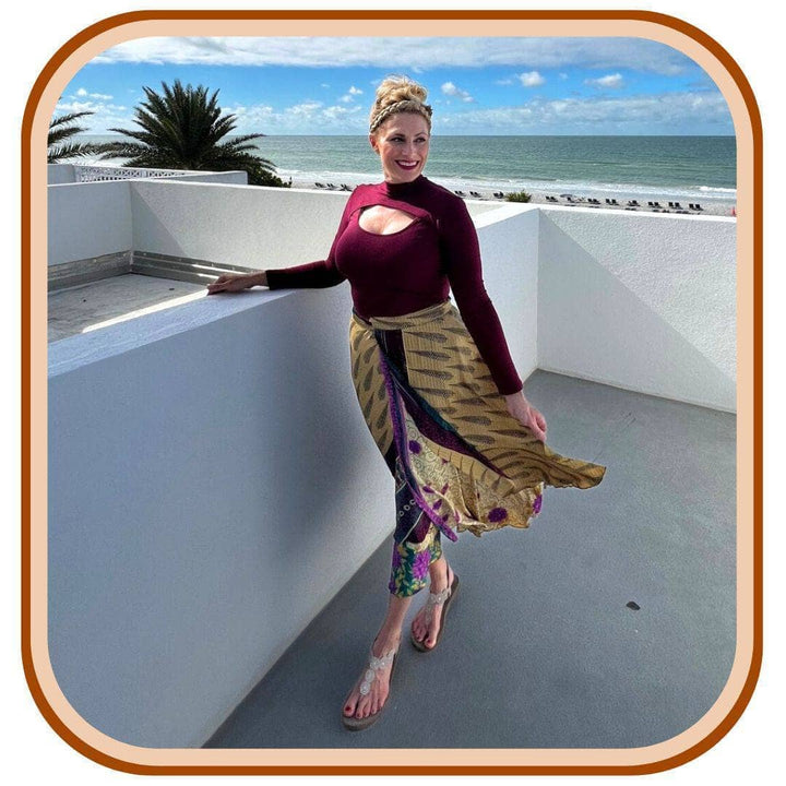 Model is standing in a tropical place. She wears a maroon long sleeve top with sandals and a sari wrap skirt. The skirt is a dusty yellow with a navy blue pattern on it. The second layer of the skirt has pink and green underneath.