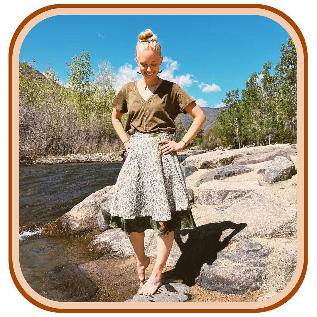Model stands on a rock with a river in the background. She wears a brown t shirt with a sari wrap skirt. The skirt is white with a dark green pattern on it. The dark green is on the second layer on the skirt.
