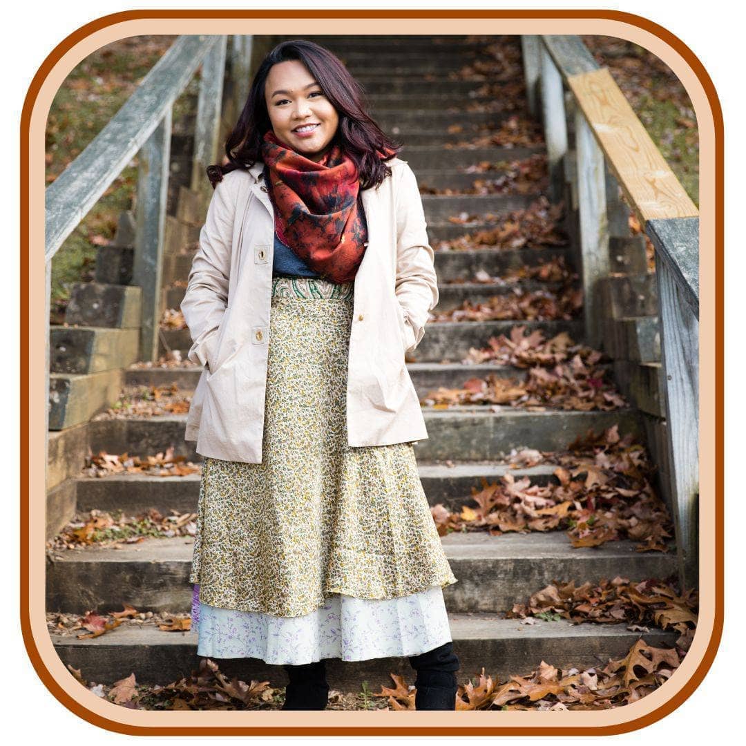 Model standing below a staircase with leaves all around her. She is wearing a Sari wrap skirt that is a dusty green and a light sky blue. She also wears a pale pink jacket, leggings and a bright red scarf.