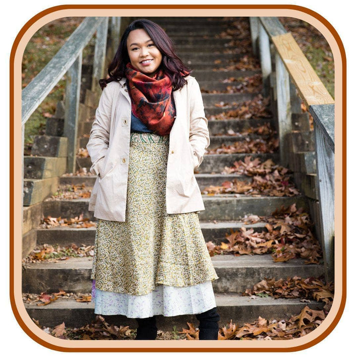 Model standing below a staircase with leaves all around her. She is wearing a Sari wrap skirt that is a dusty green and a light sky blue. She also wears a pale pink jacket, leggings and a bright red scarf.