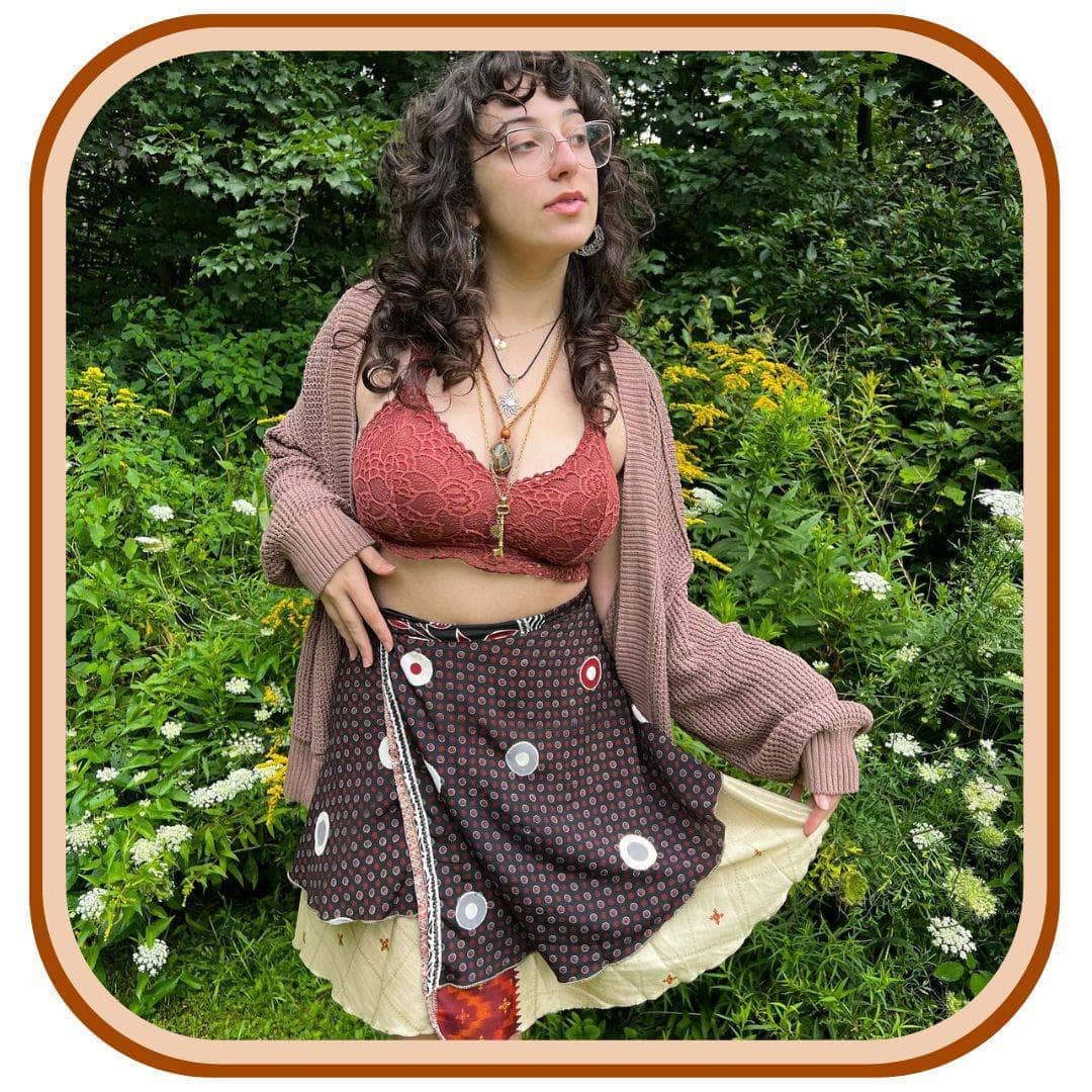 Model stands with tall grass in the background. She wears a pink crop top, with a light pink jacket, and a sari wrap skirt. The skirt is black with a dense pattern of pink and grey-pink on it. There is white dots scattered along the skirt. There is also a pale yellow trim along the bottom of the skirt.