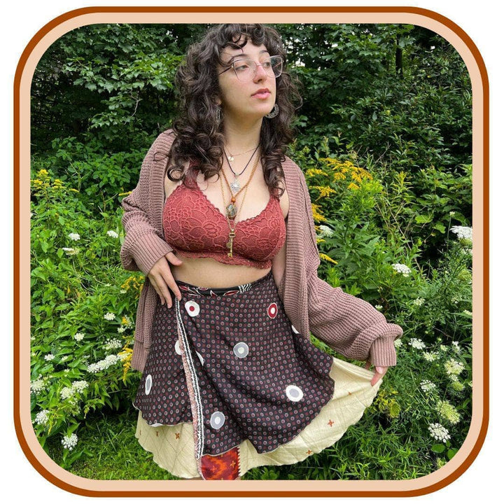 Model stands with tall grass in the background. She wears a pink crop top, with a light pink jacket, and a sari wrap skirt. The skirt is black with a dense pattern of pink and grey-pink on it. There is white dots scattered along the skirt. There is also a pale yellow trim along the bottom of the skirt.