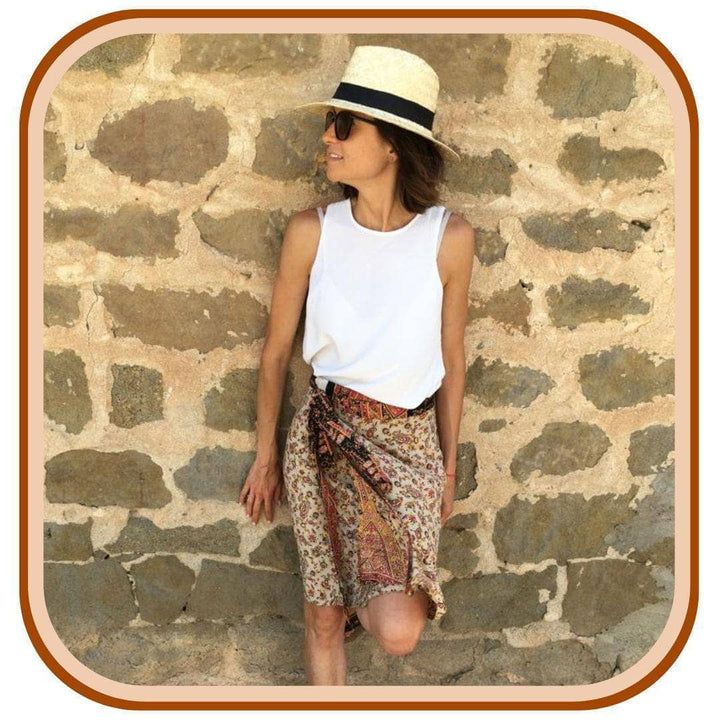 Model leans against a grey brick wall. She wears a white sun hat, a white tank top and a sari wrap skirt. The skirt is mostly white with a floral pattern throughout. The skirt has an orange belt and a dark brown second layer.