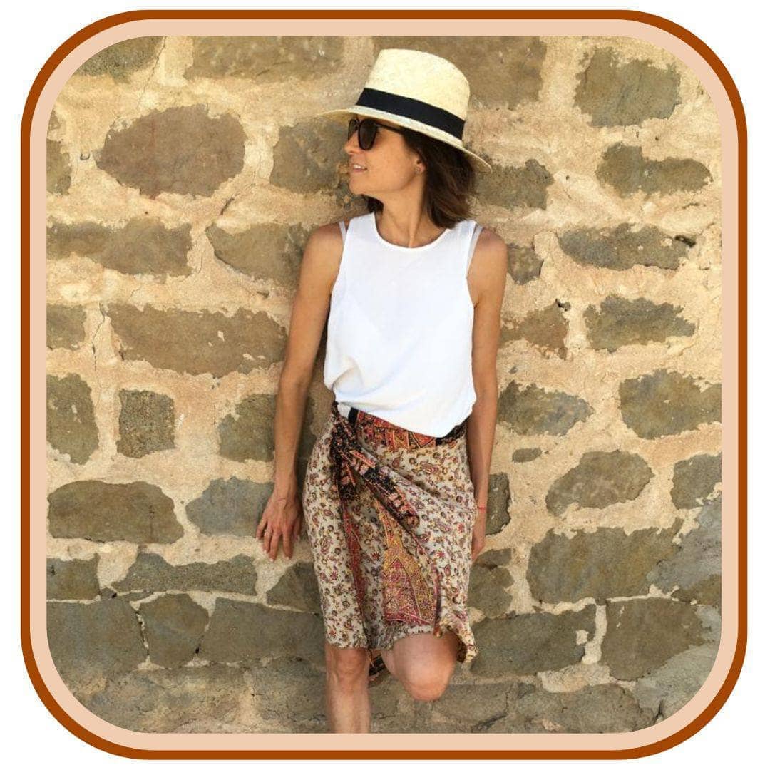 Model leans against a grey brick wall. She wears a white sun hat, a white tank top and a sari wrap skirt. The skirt is mostly white with a floral pattern throughout. The skirt has an orange belt and a dark brown second layer.