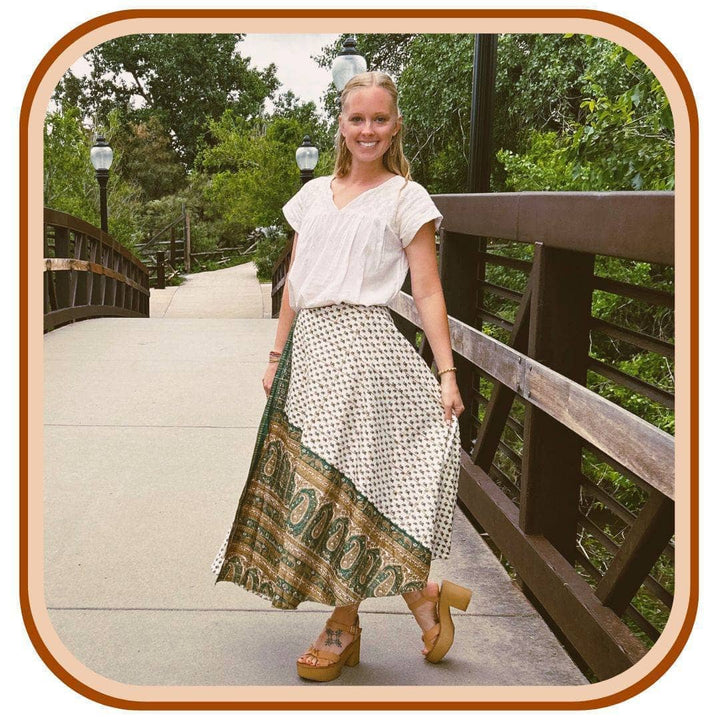 Model stands on a flowing bridge. She wears a white top and a sari wrap skirt. The skirt is mostly white with a dark green pattern.