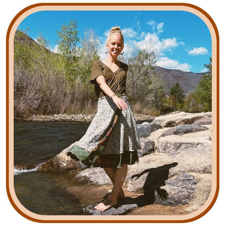 Model stands on a rock with a river in the background. She wears a brown t shirt with a sari wrap skirt. The skirt is white with a dark green pattern on it. The dark green is on the second layer on the skirt.