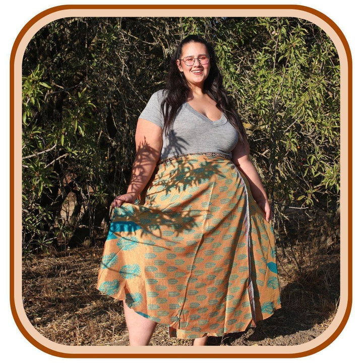 Model stands with trees in the background. She wears a light grey top with a sari wrap skirt. The skirt is a dark yellow with a turquoise pattern.