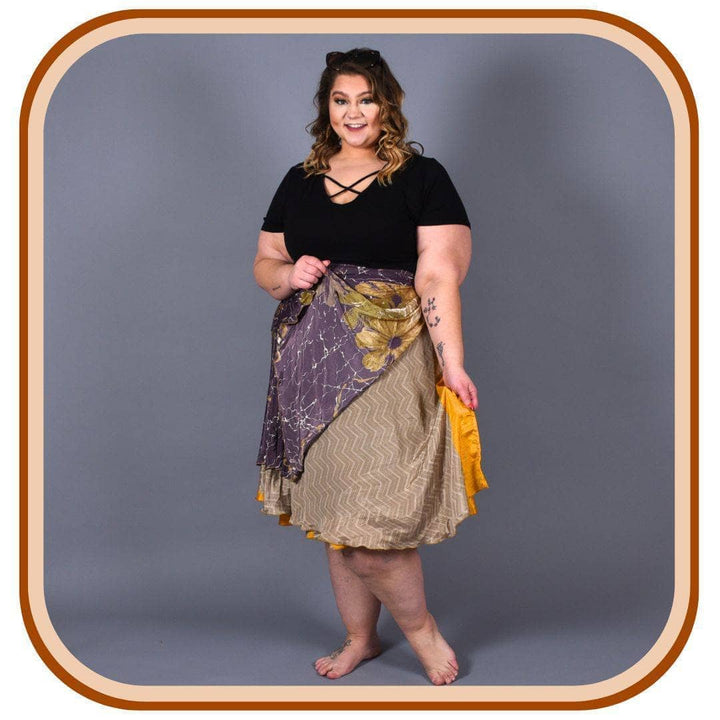 Model stands against a blank dark grey background. She wears a black top and a sari wrap skirt. The skirt is a pale purple, tan and yellow. On each layer of the skirt there is a different pattern.