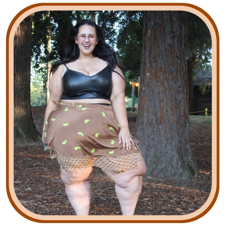 Model stands outside next to a tree. She wears a black crop top with a sari wrap skirt. The skirt is a light brown with a light green pattern on it. Below the skirt on the trim there is a lighter brown pattern.