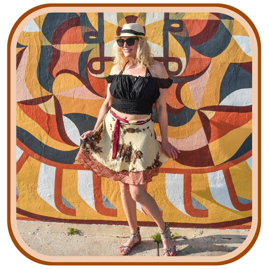 Model stands outside next to a bright mural. She wears black sunglasses a tan sun hat, a black crop top and silver sandals.  She wears a dusty yellow sari skirt with a floral pattern. The skirt has a red belt and a dark orange trim along the end of the skirt.