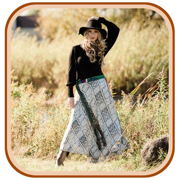 Model is standing in a dreamy golden field. She wears a black long sleeved top and a black sun hat. She wears a white sari skirt with a checkered-like pattern. The skirt has a green belt that is flowing in the wind.