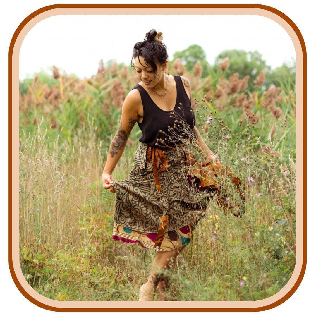 Model is in a field of tall grass. She wears a black top with a sari wrap skirt. The skirt is tan with a black pattern. The bottom trim of the skirt is a light tan with a green and maroon pattern. 