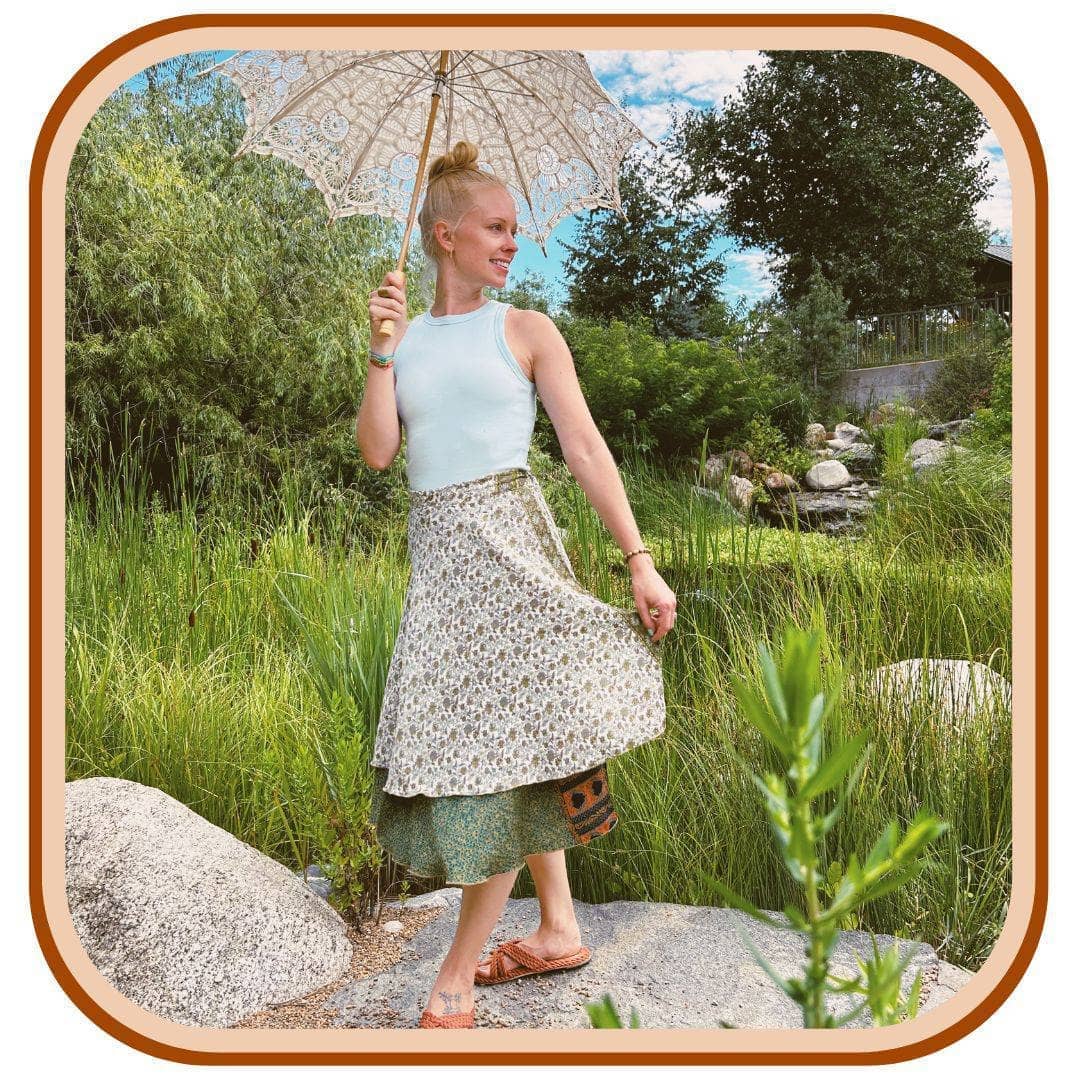 Model stands on top of a rock. She is wearing a white tank top with a sari wrap skirt.The skirt is white with a dark green pattern on it. The dark green is on the second layer on the skirt.