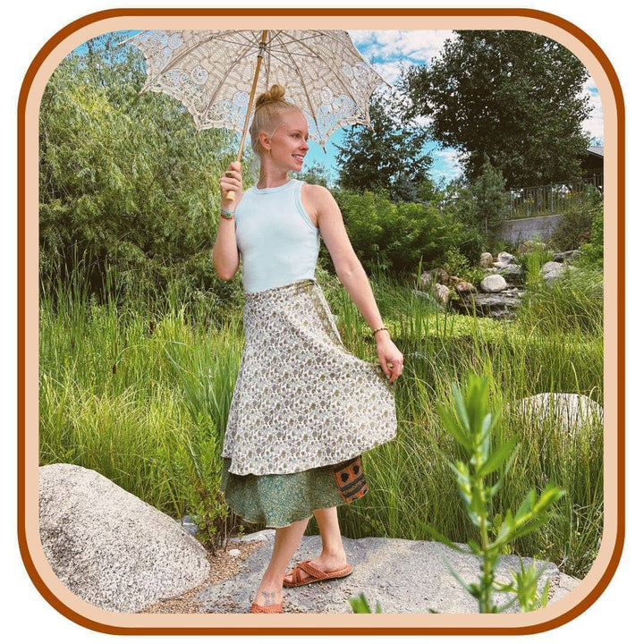 Model stands on top of a rock. She is wearing a white tank top with a sari wrap skirt.The skirt is white with a dark green pattern on it. The dark green is on the second layer on the skirt.