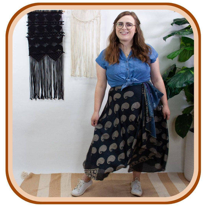 Model stands next to two macrame projects on the wall. She is wearing a denim short sleeved shirt and white sneakers. She is  wearing a sari wrap skirt that is a dark blue with one consistent pattern on the skirt.