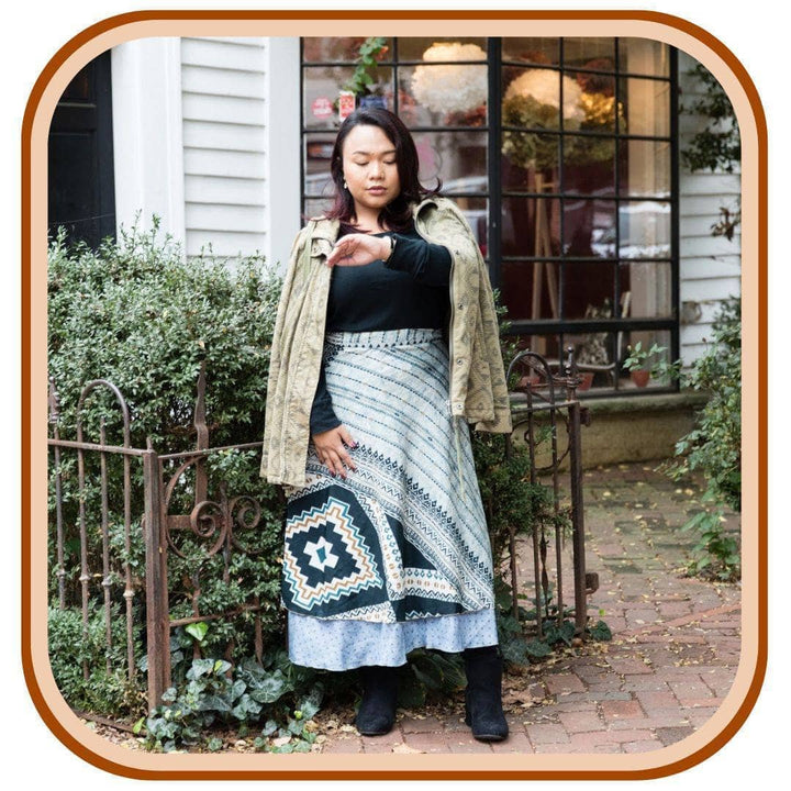 Model stands with a house in the background.She wears a light green jacket, a black long sleeve top and black boots. She wears a light blue sari wrap skirt with a striped pattern along the top of the skirt. The skirt has a sky blue.