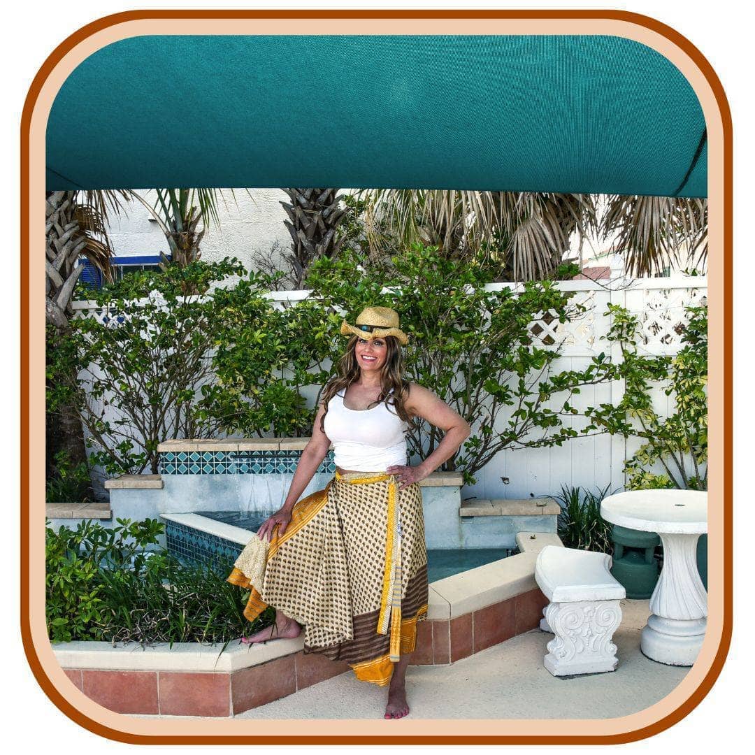Model stands outside near a tropical pool. She wears a white tank top and a tan sun hat. She wears a yellow and pale yellow sari wrap skirt. The skirt has two patterns as well as solid colors.