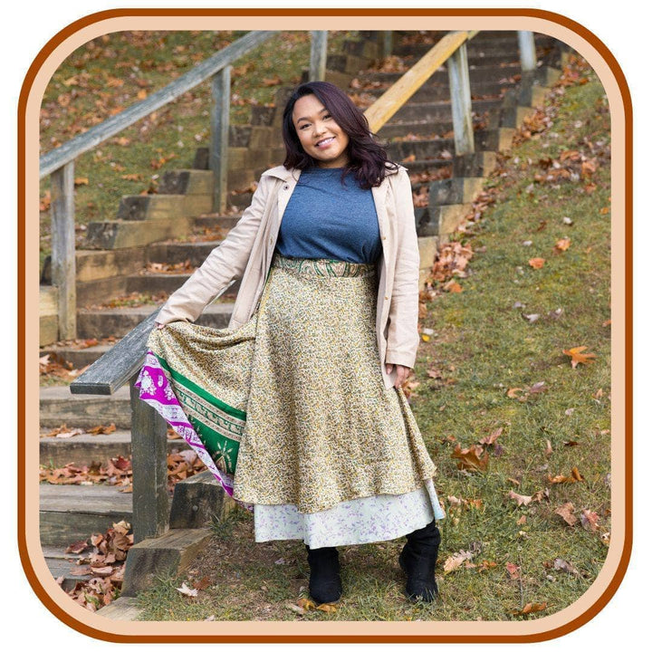 Model standing below a staircase with leaves all around her. She is wearing a Sari wrap skirt that is a dusty green and a light sky blue with a green and pink pattern underneath the skirt. She also wears a pale pink jacket, leggings and a blue t shirt.