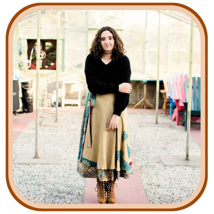 Model stands in a sunroom. She wears a black long sleeve top, tan boots, and a sari wrap skirt. The skirt is tan with a turquoise pattern on it.