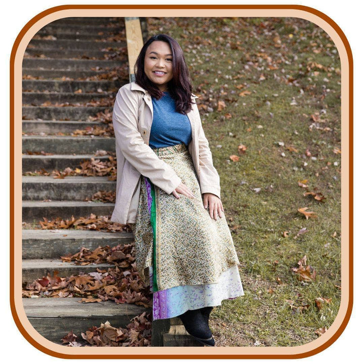 Model standing below a staircase with leaves all around her. She is wearing a Sari wrap skirt that is a dusty green and a light sky blue with a green and pink pattern underneath the skirt. She also wears a pale pink jacket, leggings and a blue t shirt.