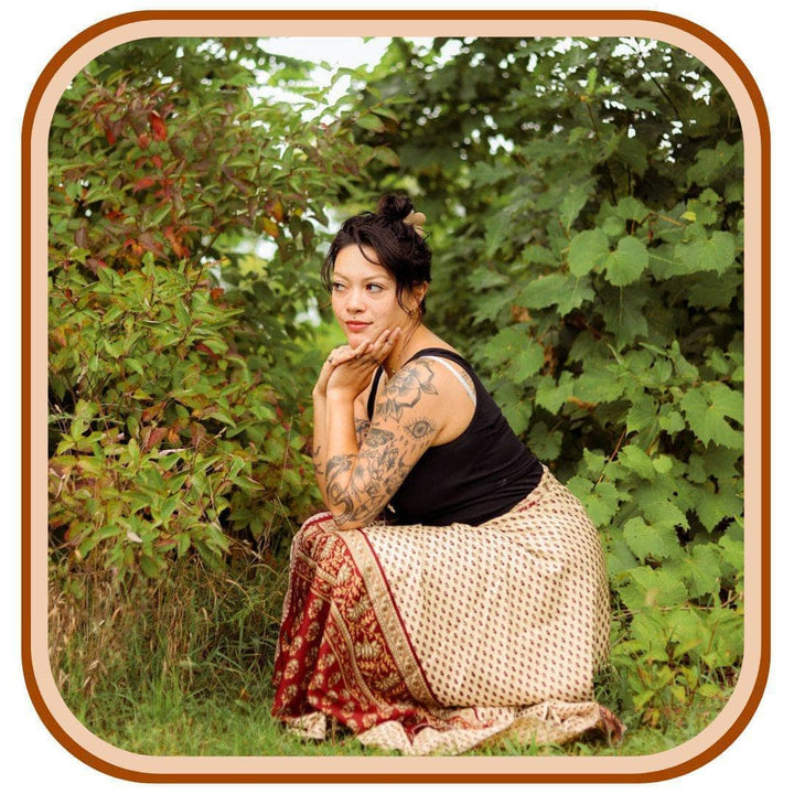 Model stands outside with the green trees. She wears a black tank top and a tan and red sari wrap skirt. The skirt features 3 patterns on each section of color.