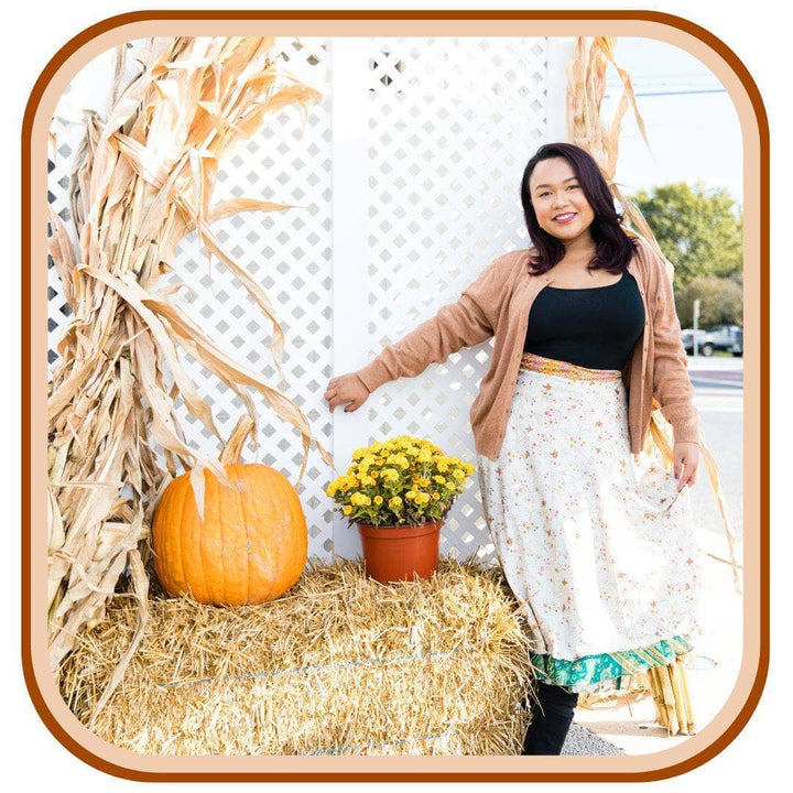 Model leans against hay bails with pumpkins on them. She wears a light brown jacket, a black top, black boots, and a sari wrap skirt. The skirt is white with a dark orange floral pattern that is very minimal. The skirt has a green trim on the bottom and an orange trim on the top of the skirt.
