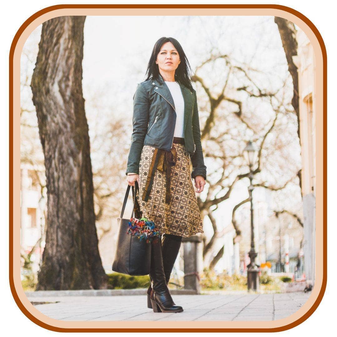 Model stands outside with trees all around her. She wears a green jacket with a white top, black boots, a black hand bag, and a sari wrap skirt. The skirt is a light tan with a black minimal pattern that looks checkered. The skirt has a brown belt.