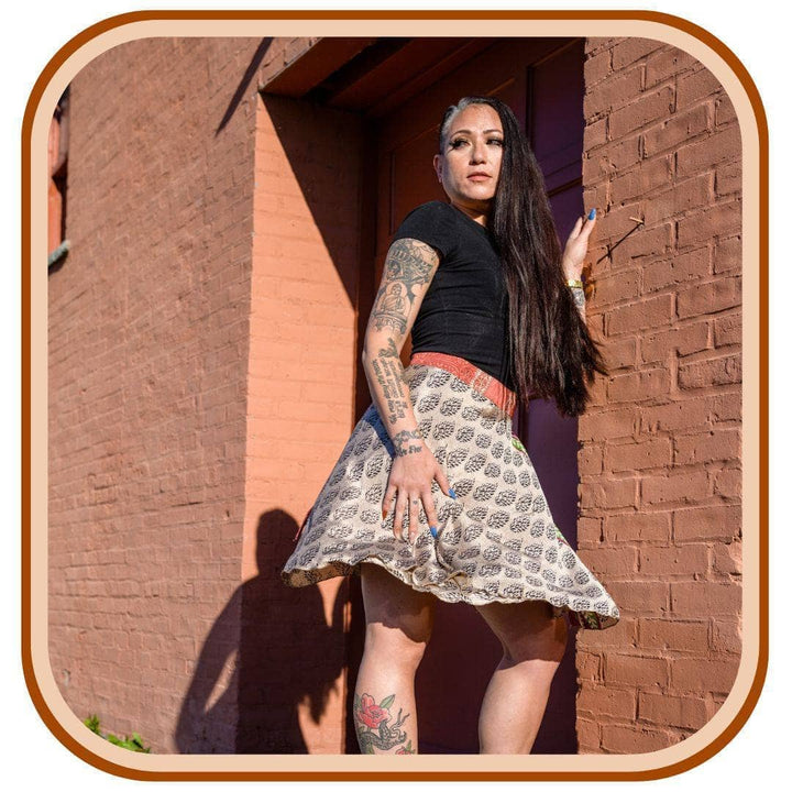 Model is leaning against a brick wall. She wears a black t shirt and a sari wrap skirt. The skirt is a dusty tan with a black pattern that is consistent throughout.