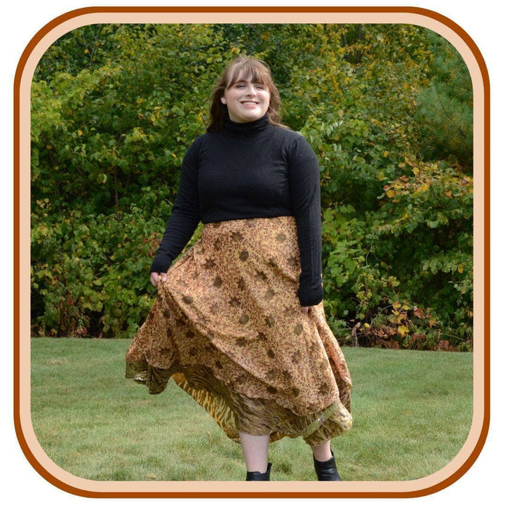 Model stands outside with trees in the background. She wears a black long sleeve top and a sari wrap skirt. The skirt is a light tan with a dark brown pattern.