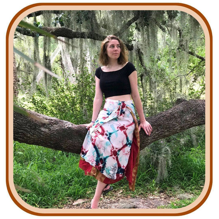 Model sits on a giant tree branch in the forest. She wears a black crop top and a sari wrap skirt. The skirt is mostly white with a turquoise and red pattern on it. 