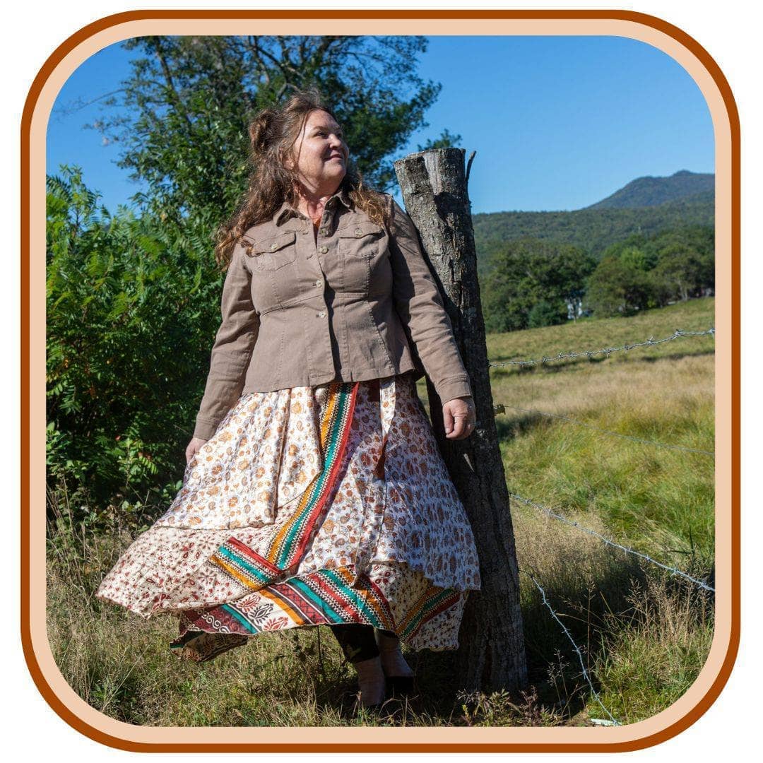 Model is outside with trees in the background. She wears a light grey jacket, and a sari wrap skirt. The skirt is mostly white with yellow flowers as a pattern. There is a yellow, green, and maroon stripe that goes through the second layer of the skirt.