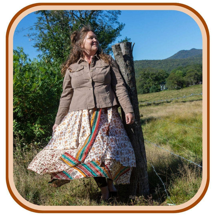 Model is outside with trees in the background. She wears a light grey jacket, and a sari wrap skirt. The skirt is mostly white with yellow flowers as a pattern. There is a yellow, green, and maroon stripe that goes through the second layer of the skirt.
