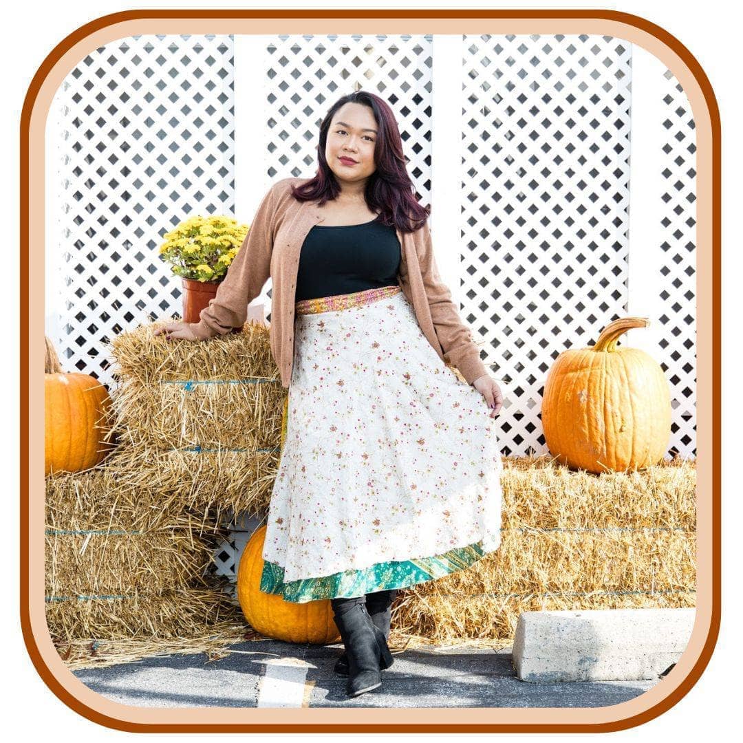 Model leans against hay bails with pumpkins on them. She wears a light brown jacket, a black top, black boots, and a sari wrap skirt. The skirt is white with a dark orange floral pattern that is very minimal. The skirt has a green trim on the bottom and an orange trim on the top of the skirt.