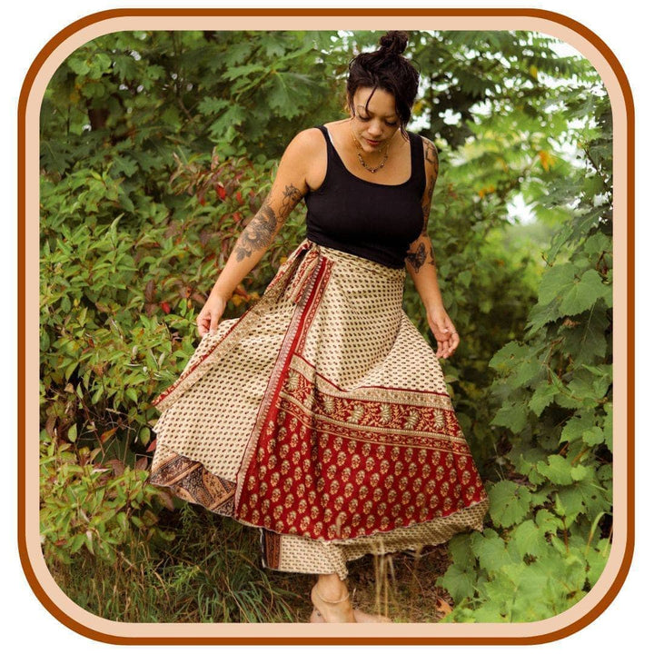 Model stands outside with the green trees.  She wears a black tank top and a  tan and red sari wrap skirt. The skirt features 3 patterns on each section of color.