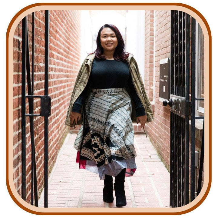Model walks through a gateway. She wears a light green jacket, a black long sleeve top and black boots. She wears a light blue sari wrap skirt with a striped pattern along the top of the skirt. The skirt has a sky blue and pink pattern underneath.